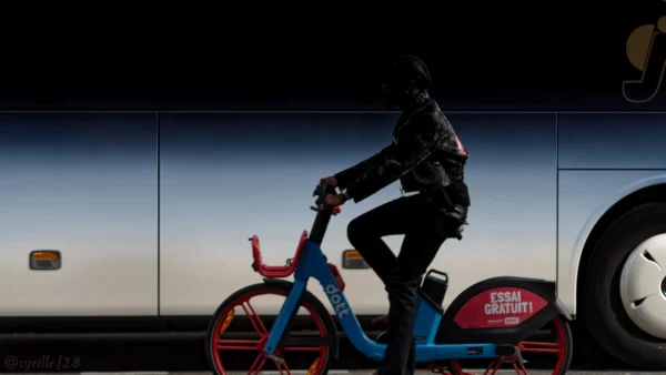 Cyrille GONTIER · 07/03/2025 · 2026 x 1140 Cette photographie met en scène un cycliste en tenue sombre, assis sur un vélo électrique bleu vif, probablement un vélo en libre-service. La composition est dynamique, avec un cadrage serré sur le sujet, isolé devant un fond flou de bus et de mur.La lumière, douce et tamisée, crée des contrastes entre ombres et clartés, accentuant le côté dramatique. Les couleurs dominantes sont les tons bleus du vélo et les teintes sombres du cycliste, avec des touches de rouge et de blanc en arrière-plan.Le style est épuré et moderne, évoquant une ambiance urbaine nocturne ou crépusculaire, à la fois calme et légèrement mystérieuse.