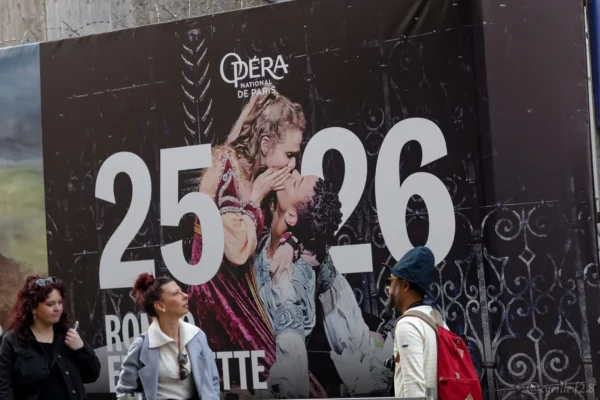 Cyrille GONTIER · 07/03/2025 · 2026 x 1351 Cette photographie montre une affiche publicitaire géante pour le film "Camerà" avec les acteurs dans les rôles de Roméo et Juliette (25-26). Le sujet principal est l’affiche elle-même, avec deux personnages stylisés en noir et blanc.La composition met en avant l’affiche centrale, encadrée par des passants en bas. Le cadrage est serré sur l’affiche et les personnes, créant un contraste entre l’artifice du film et la réalité. La lumière est naturelle, avec des tons clairs et contrastés. Les couleurs dominantes sont le noir, blanc et rouge. L’ambiance est urbaine et cinématographique.