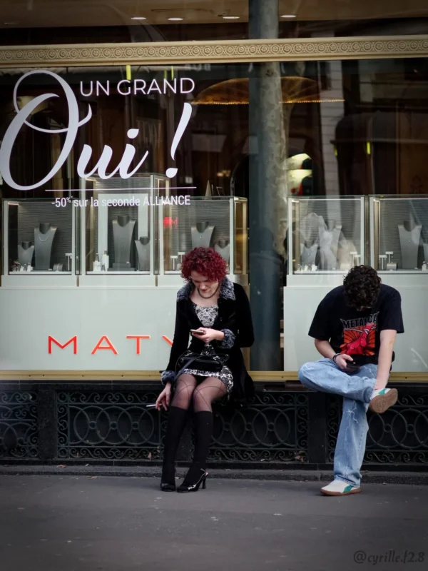 Cyrille GONTIER · 07/03/2025 · 1520 x 2026 Cette photographie capture deux personnes assises sur un trottoir devant une vitrine. Le sujet principal est la femme à gauche, aux cheveux roses, vêtue de noir et utilisant un téléphone, tandis que l’homme à droite, en jeans et veste, semble détendu.La composition est symétrique et équilibrée, avec un cadrage serré sur les deux individus. La lumière est naturelle, douce, avec des ombres marquées. Les couleurs dominantes sont les tons froids (noir, gris) et quelques touches chaudes (rouge de la veste de l’homme, cheveux roses).Le style est réaliste et documentaire, évoquant une ambiance urbaine et décontractée.