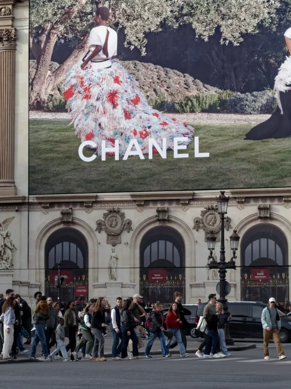 Cyrille GONTIER · 07/03/2025 · 1520 x 2026 Cette photographie montre une façade de magasin **Chanel** avec une grande enseigne représentant une silhouette féminine stylisée, vêtue d’un manteau à motifs floraux rouges et blancs. Le **sujet principal** est cette affiche emblématique.La **composition** met en valeur l’architecture classique du bâtiment, avec ses colonnes et ses détails ornementaux. Le **cadrage** inclut une foule de passants, créant un contraste entre le luxe et l’agitation urbaine. La **lumière** semble naturelle, avec des ombres douces. Les **couleurs dominantes** sont les tons neutres de la pierre, rehaussés par les rouges et blancs de l’affiche.Le **style** est élégant et sophistiqué, reflétant le prestige de la marque. L’**ambiance** est dynamique, mêlant raffinement et vie quotidienne parisienne.