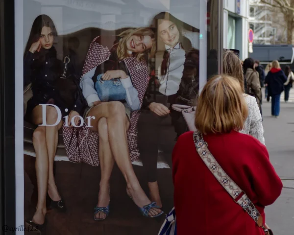 Cyrille GONTIER · 07/03/2025 · 2026 x 1620 Cette photographie montre une vitrine Dior reflétant trois mannequins stylisés, vêtus de tenues élégantes et décontractées. Le sujet principal est la réflexion des mannequins, contrastant avec une femme en rouge à l'avant-plan, portant un badge.La composition utilise un cadrage serré sur la vitrine, capturant aussi les passants floutés en arrière-plan. La lumière est naturelle, probablement diurne, avec des ombres douces. Les couleurs dominantes sont les tons froids (bleu, gris) et chauds (rouge vif du manteau de la femme).L’ambiance est urbaine et chic, mêlant élégance et vie quotidienne. Le style est photographique, réaliste et légèrement artistique grâce aux jeux de réflexion.