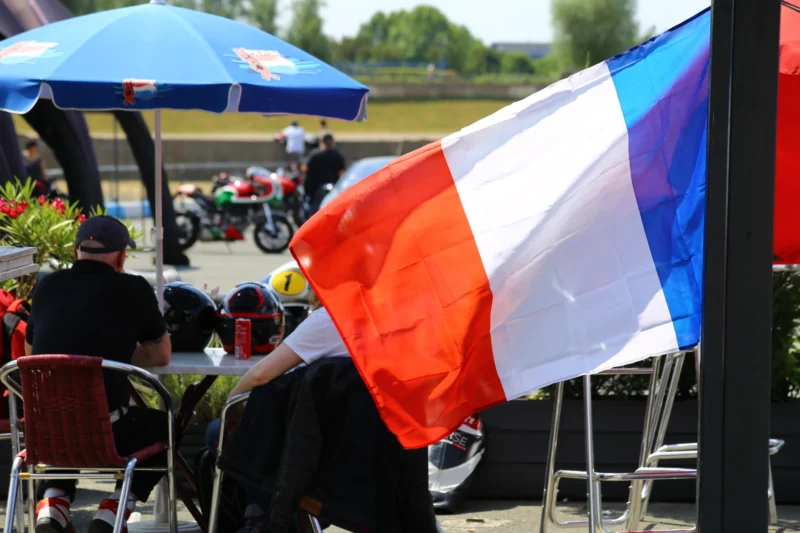 Frédéric · 15/07/2018 · 5472 x 3648 Cette photographie met en scène un drapeau français en premier plan, agité par le vent. La composition montre des spectateurs assis sous un parasol bleu, en arrière-plan, avec des motos et des personnes en mouvement.Le cadrage est serré sur le drapeau, mais inclut aussi des éléments contextuels. La lumière est naturelle, probablement en journée, avec des ombres douces. Les couleurs dominantes sont le bleu, blanc, rouge du drapeau, et le bleu du parasol.L’ambiance évoque une scène informelle, peut-être un événement sportif ou une rassemblement, avec une touche patriotique. Le style est documentaire et spontané.