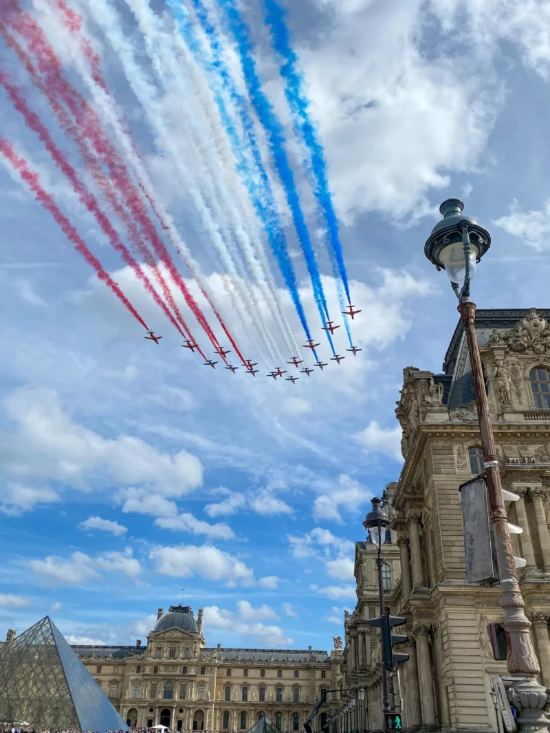 Daniel · 2681 x 3575 Cette photographie capture un spectacle aérien avec des avions traçant des fumées colorées (bleu, blanc, rouge) dans le ciel. Le sujet principal est cette formation en vol, symbolisant souvent une célébration patriotique.La composition montre en premier plan un bâtiment historique parisien, probablement le Louvre, avec une pyramide visible. Le cadrage est dynamique, intégrant à la fois le ciel et l’architecture.La lumière est naturelle, avec un ciel partiellement nuageux. Les couleurs dominantes sont les teintes vives des traînées et les tons gris-beige des bâtiments. L’ambiance est festive et majestueuse, mêlant tradition et modernité.