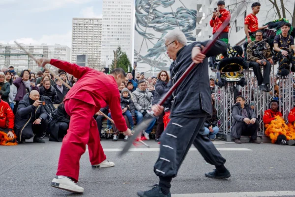 Frédéric · 2517 x 1680 Cette photographie capture deux hommes en pleine démonstration de combat à l'épée sur une place publique. Le sujet principal est leur duel dynamique, encadré en plan serré.La composition montre un premier plan centré sur les combattants, avec en arrière-plan une foule et des bâtiments urbains. La lumière est naturelle, probablement diurne, avec des ombres marquées. Les couleurs dominantes sont les tons vifs du costume rouge d'un combattant et les gris/bleus du vêtement de l'autre, contrastant avec les teintes neutres de l'environnement.L’ambiance est à la fois traditionnelle et moderne, mêlant spectacle et vie urbaine.