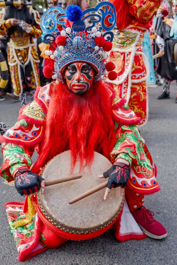 Frédéric · 1121 x 1680 Cette photographie met en scène un personnage vêtu d’un costume traditionnel richement coloré, probablement lié à une célébration culturelle asiatique. Le sujet principal, un homme masqué et paré de rouge et d’or, joue d’un tambour.La composition est centrée sur lui, avec un cadrage serré qui isole le personnage. Les couleurs dominantes sont le rouge éclatant, l’or et les teintes vives des motifs. La lumière semble naturelle et met en valeur les détails du costume. L’ambiance est festive et théâtrale, évoquant une tradition vivante et une célébration culturelle.
