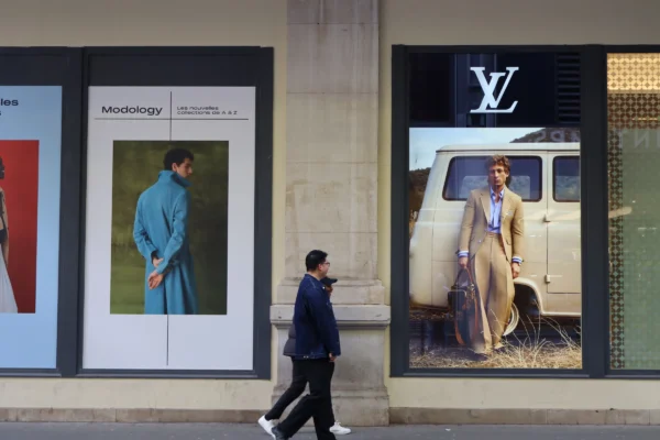 Camilla · 07/03/2026 · 6000 x 4000 Cette photographie montre un homme marchant devant une vitrine Louis Vuitton. Le sujet principal est la vitrine avec deux affiches publicitaires : une femme en tenue élégante et un homme dans un véhicule vintage. La composition est symétrique, avec un cadrage serré sur la vitrine et l’homme en mouvement. La lumière est naturelle et douce, éclairant les visages et les détails des affiches. Les couleurs dominantes sont les tons neutres (beige, gris) et les touches de vert et bleu. Le style est urbain et sophistiqué, évoquant une ambiance chic et moderne.