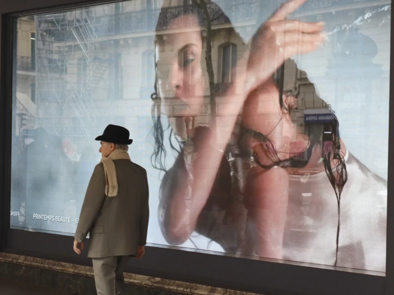 Cette photographie montre un homme en costume et chapeau, dos tourné, observant une projection géante derrière lui. Le sujet principal est cette projection représentant une femme en tenue futuriste, brandissant un objet.La composition oppose l’homme discret et la scène dynamique projetée. Le cadrage est serré sur l’homme, avec une profondeur de champ qui met en valeur l’écran lumineux. La lumière est artificielle et contrastée, éclairant fortement la projection.Les couleurs dominantes sont froides et métalliques, avec des teintes grises et bleutées. Le style semble inspiré par un univers cyberpunk ou science-fiction. L’ambiance est à la fois mystérieuse et immersive.