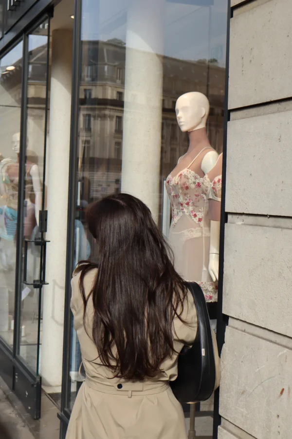 Camilla · 07/03/2026 · 3704 x 5557 Cette photographie montre une femme vue de dos, observant une mannequin vêtue d’une robe à fleurs dans une vitrine. Le sujet principal est cette interaction entre la femme et la mannequin.La composition est simple et centrée, avec un cadrage serré sur les deux figures. La lumière est naturelle et douce, éclairant les visages et les détails des vêtements. Les couleurs dominantes sont neutres (beige, gris) avec des touches vives apportées par la robe fleurie.L’ambiance est urbaine et contemplative, évoquant une pause dans une rue commerçante. Le style est réaliste et quotidien.