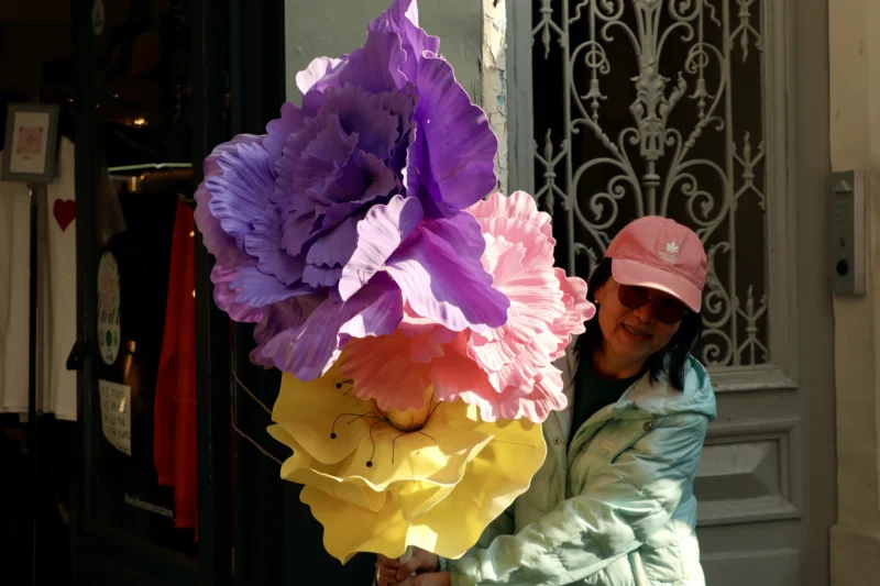 Francoise · 21/03/2026 · 3840 x 2560 Cette photographie montre une personne portant un immense chapeau coloré en forme de fleur, composé de pétales violets, roses et jaunes. Le sujet principal est donc ce chapeau spectaculaire.La composition est simple et centrée sur la personne, avec un cadrage serré qui met en valeur les détails du chapeau. La lumière semble naturelle, bien que légèrement tamisée, créant des ombres douces. Les couleurs dominantes sont vives et pastel, apportant une touche joyeuse et festive.Le style est photographique et spontané, évoquant une ambiance artistique et folklorique.