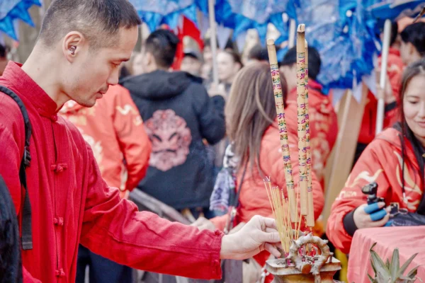 Frédéric · 2520 x 1680 Cette photographie capture une scène traditionnelle où le sujet principal est un homme en rouge allumant des bâtons d’encens. La composition montre plusieurs personnes en arrière-plan, légèrement floues, créant une ambiance animée. Le cadrage est serré sur les mains de l’homme et les bâtons d’encens, avec une profondeur de champ réduite.La lumière est douce et naturelle, mettant en valeur les détails. Les couleurs dominantes sont les rouges vifs, les bleus et les tons chauds des vêtements et accessoires. Le style est documentaire et spontané, évoquant une ambiance culturelle et spirituelle, typique d’un lieu de culte ou de célébration.