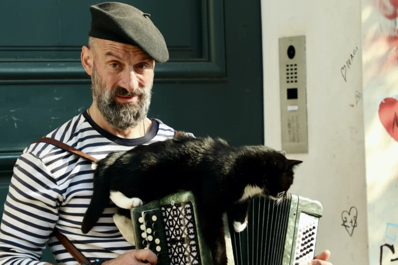 Francoise · 21/03/2026 · 3840 x 2560 Cette photographie met en scène un homme comme sujet principal, vêtu d’un pull à rayures et d’un béret, jouant d’un accordéon. Deux chats noirs sont posés sur l’instrument.La composition est serrée, avec un cadrage en plan moyen. La lumière semble naturelle et douce, éclairant le sujet sans ombres marquées. Les couleurs dominantes sont les tons neutres (gris, noir) et les rayures blanches du pull.Le style est documentaire et spontané, évoquant une ambiance chaleureuse et intimiste, presque poétique.