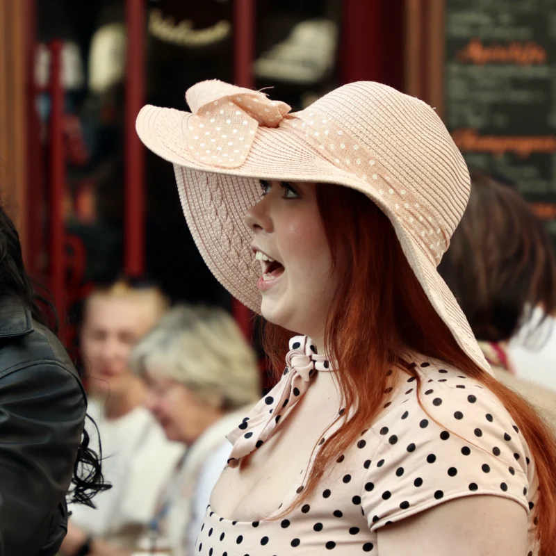 Francoise · 21/03/2026 · 3840 x 3840 Cette photographie met en scène une femme en hat de paille à larges bords et vêtue d’une robe à pois, comme sujet principal. Le cadrage est serré sur son visage, légèrement flou, créant un effet de profondeur.La lumière semble naturelle, douce et légèrement tamisée, mettant en valeur les tons chauds. Les couleurs dominantes sont les teintes pastel (blanc, beige) et les touches de rouge dans les cheveux. L’ambiance évoque une scène rétro ou vintage, peut-être inspirée d’une époque ancienne ou d’un événement théâtral. L’arrière-plan flou et les autres personnes suggèrent une foule ou un rassemblement.