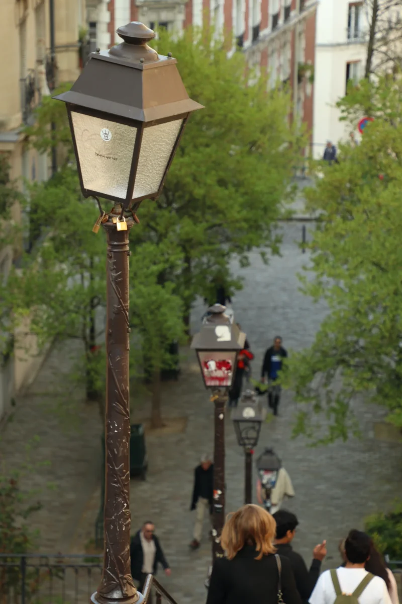 Francoise · 21/03/2026 · 3840 x 5760 Cette photographie montre une rue pavée en pente, avec une **lampe à gaz ornée** comme sujet principal. La **composition** est dynamique, avec des lignes convergentes vers le haut de la rue.Le **cadrage** est légèrement incliné, offrant une perspective immersive. La **lumière** est naturelle, douce, avec des ombres légères, suggérant un jour nuageux. Les **couleurs dominantes** sont des tons neutres (gris, beige) rehaussés par le vert des arbres et le rouge de la lampe.Le style évoque un **ambiance romantique et pittoresque**, typique d’un quartier historique européen, mêlant charme ancien et vie quotidienne.