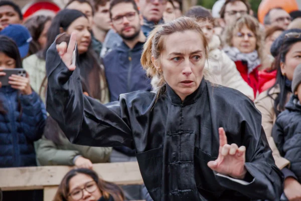Frédéric · 2517 x 1680 Cette photographie met en scène une femme comme sujet principal, vêtue d’un manteau noir, faisant un geste de la main. Elle semble être au centre d’une foule attentive, en extérieur.La composition est dynamique, avec un cadrage serré sur la femme, entourée d’un public en arrière-plan. La lumière est naturelle, légèrement tamisée, créant des contrastes doux. Les couleurs dominantes sont les tons sombres (noir, gris) et quelques touches de couleurs chaudes (vêtements du public).Le style est réaliste et cinématographique, évoquant une ambiance théâtrale ou une scène de film. L’atmosphère est intense et captivante.