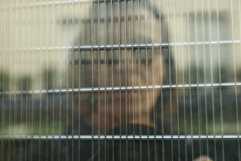 Francoise · 21/03/2026 · 3840 x 2559 Cette photographie montre un visage flou vu à travers une structure métallique en treillis, probablement une cage ou une grille. Le sujet principal est indistinct, ce qui crée une impression de mystère.La composition est simple, centrée sur le visage partiellement caché. Le cadrage serré et les lignes horizontales de la grille dominent l'image, ajoutant une sensation de confinement.Les couleurs sont douces et neutres, avec des teintes grises et bleutées. La lumière semble naturelle mais tamisée, renforçant l'ambiance mélancolique et introspective. Le style est épuré et minimaliste, évoquant une atmosphère contemplative et légèrement mélancolique.