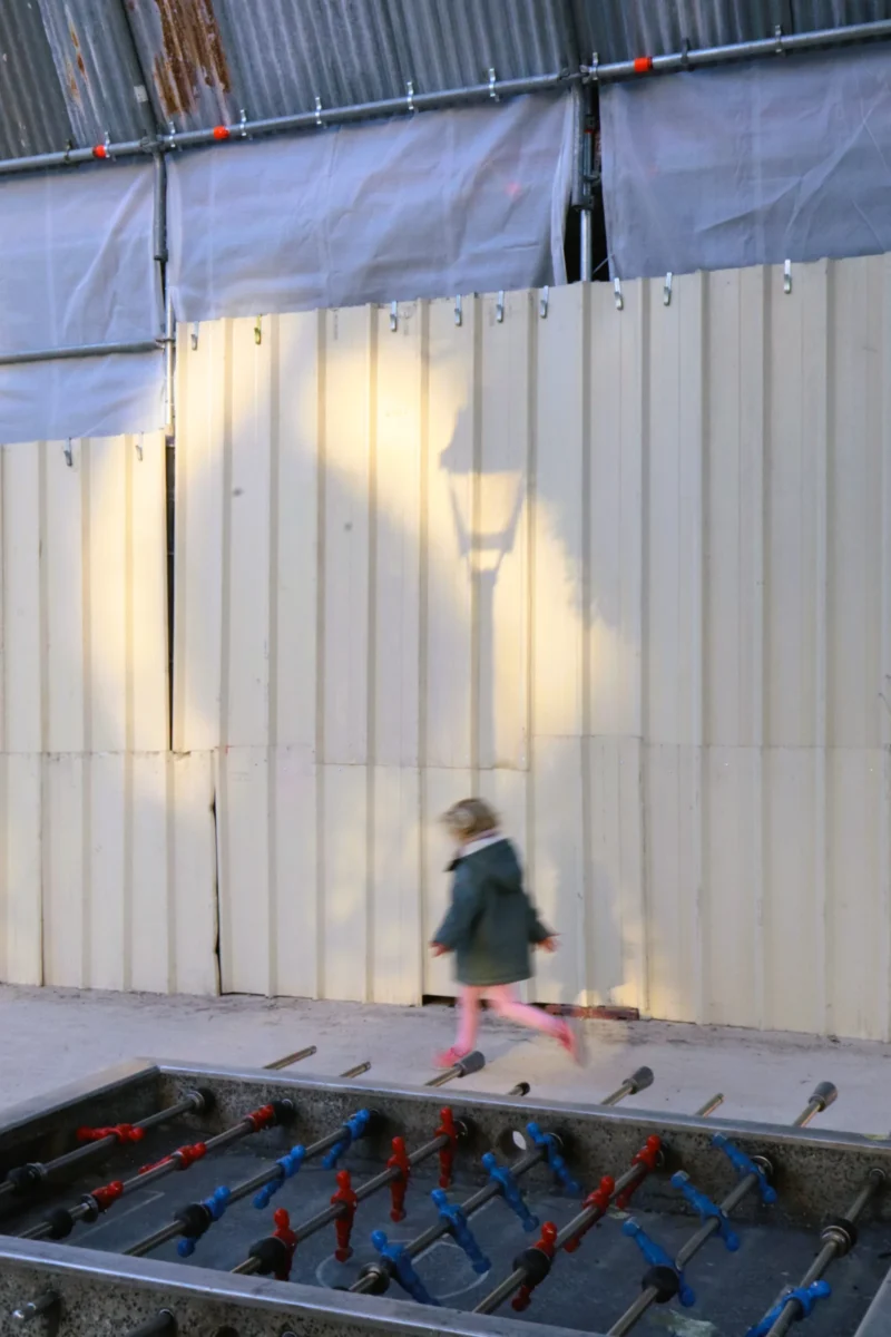 Camilla · 4000 x 6000 Cette photographie capture une enfant en mouvement devant une structure métallique et des containers blancs. Le sujet principal est la fillette, vêtue d’une robe sombre, dont les pieds ne touchent presque pas le sol, suggérant une course ou un saut.La composition est dynamique, avec un cadrage serré sur le bas du corps. La lumière naturelle, douce et légèrement dorée, traverse les interstices des containers, créant des jeux d’ombres et de clarté. Les couleurs dominantes sont les tons gris métalliques, blancs et quelques touches de rouge (les chaussures). L’ambiance est à la fois ludique et poétique, évoquant liberté et insouciance.