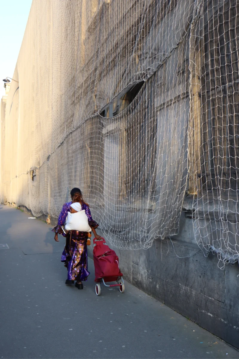 Camilla · 3825 x 5738 Cette photographie montre deux enfants marchant côte à côte sur un trottoir. Le sujet principal est l’enfant de dos, vêtu d’un t-shirt blanc, poussant un vélo avec une remorque rouge contenant un autre enfant.La composition est simple et horizontale, avec un cadrage serré sur les enfants. La lumière semble naturelle, douce et légèrement tamisée, créant des ombres discrètes. Les couleurs dominantes sont les tons neutres (gris du mur, beige du sol) et les touches de rouge et blanc des vêtements.L’ambiance est calme et quotidienne, évoquant un moment de vie simple et authentique.