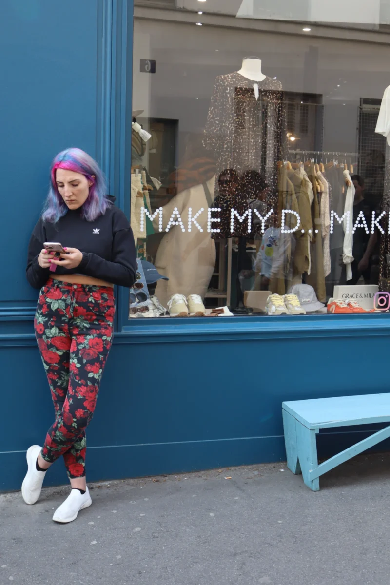Camilla · 4000 x 6000 Cette photographie montre une personne, sujet principal, vêtue d’un bonnet coloré et de pantalons à motifs, absorbée par son téléphone. Elle est positionnée devant une vitrine bleue avec des mannequins et des accessoires.Le cadrage est serré sur elle, avec une composition équilibrée entre le sujet et l’arrière-plan. La lumière semble naturelle, légèrement diffuse, mettant en valeur les détails des vêtements et de la vitrine. Les couleurs dominantes sont vives (rose, bleu, motifs variés) créant un contraste dynamique.L’ambiance est urbaine et décontractée, avec une touche artistique et moderne.