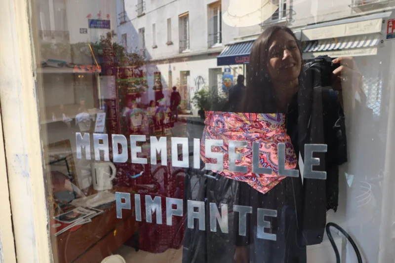 Camilla · 21/03/2026 · 6000 x 4000 Cette photographie montre une personne derrière une vitrine tenant un panneau avec l'inscription "Mademoiselle Pimpante". Le sujet principal est cette personne, partiellement cachée par le cadre.La composition est simple, avec un cadrage serré sur la vitrine et ses éléments décoratifs. La lumière semble naturelle, éclairant le sujet de manière douce. Les couleurs dominantes sont des tons chauds et pastel, avec des touches de rouge et de blanc.Le style est réaliste et spontané, évoquant une ambiance artisanale et chaleureuse, typique d’un petit commerce ou d’une boutique vintage.
