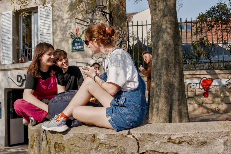 Frédéric · 21/03/2026 · 2517 x 1680 Cette photographie montre trois jeunes femmes assises sur un muret en pierre. Le sujet principal est leur interaction détendue, avec une ambiance conviviale et informelle.La composition est naturelle, avec un cadrage serré sur les trois personnes. La lumière est douce et naturelle, probablement en journée, éclairant les visages et créant des ombres légères. Les couleurs dominantes sont des tons vifs : rouge, bleu, vert et blanc.Le style est réaliste et spontané, évoquant une scène de vie quotidienne. L’ambiance est chaleureuse et décontractée, renforcée par les expressions et postures des jeunes femmes.