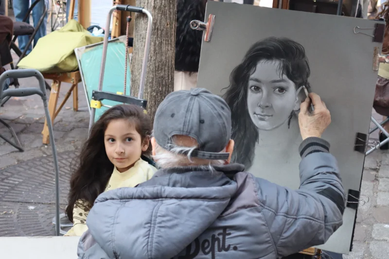 Camilla · 21/03/2026 · 6000 x 4000 Cette photographie montre une jeune fille en premier plan, vêtue d’un manteau gris et d’un bonnet, regardant une grande affiche publicitaire derrière elle. Le sujet principal est cette affiche représentant une femme stylisée, aux traits doux et modernes.La composition juxtapose l’enfant et l’affiche, créant un contraste entre le naturel et l’artistique. Le cadrage est serré sur les deux sujets. La lumière semble naturelle, avec des ombres douces. Les couleurs dominantes sont les tons froids (gris, bleu) et quelques touches chaudes (jaune du siège).L’ambiance est à la fois urbaine et intime, mêlant quotidien et art.