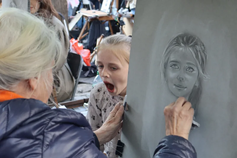 Camilla · 21/03/2026 · 6000 x 4000 Cette photographie montre une scène de portrait en train d’être dessiné. Le sujet principal est une jeune fille observant son portrait en cours de réalisation sur une ardoise.La composition est serrée, mettant en valeur les interactions entre les personnes. Le cadrage est rapproché, centré sur les visages et les mains. La lumière semble naturelle, douce et légèrement tamisée. Les couleurs dominantes sont les tons froids (gris, bleu) et quelques touches chaudes (orange du vêtement).L’ambiance est conviviale et artistique, évoquant un moment de création et de partage.
