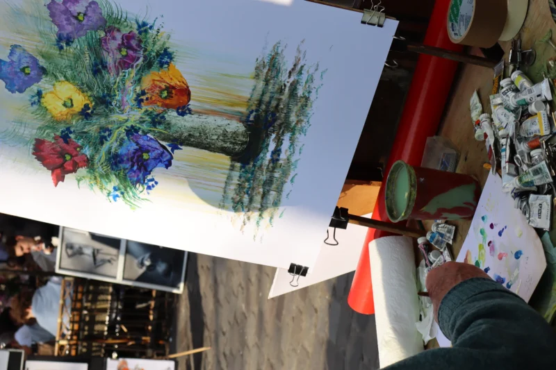 Camilla · 21/03/2026 · 6000 x 4000 Cette photographie montre une personne tenant une grande toile colorée représentant un bouquet de fleurs stylisées et un paysage. Le sujet principal est cette peinture aux couleurs vives, dominées par des teintes bleues, violettes et vertes.La composition est dynamique, avec un cadrage serré sur la toile, mettant en valeur ses détails. La lumière semble naturelle, bien répartie, éclairant les couleurs sans ombre marquée. L’ambiance est artistique et créative, évoquant un marché ou une exposition d’art. Le style semble naïf ou artisanal, avec des traits expressifs et des couleurs saturées.