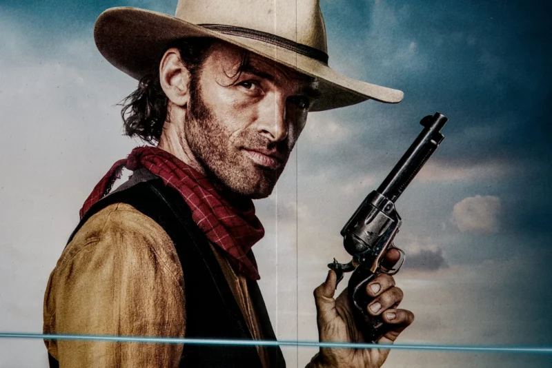 Frédéric · 21/03/2026 · 2517 x 1680 Cette photographie met en scène un homme incarnant un cow-boy, sujet principal. Il porte un chapeau, une veste en cuir et tient un revolver, évoquant un style western. La composition est centrée sur son visage et son arme, avec un cadrage serré. La lumière est naturelle et contrastée, mettant en valeur son expression déterminée. Les couleurs dominantes sont les tons chauds (beige, marron) et le rouge de sa veste, contrastant avec le ciel bleu clair. L’ambiance est dramatique et nostalgique, typique des films de western.