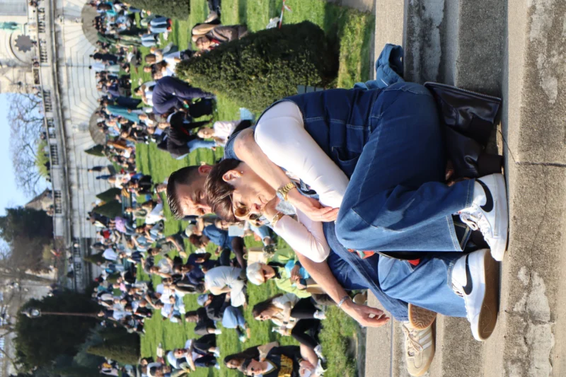 Camilla · 21/03/2026 · 6000 x 4000 Cette photographie montre deux personnes assises sur une corniche, dominant une foule en contrebas. Le sujet principal est le couple, dont l’un semble utiliser un appareil photo ou un téléphone.La composition est dynamique, avec un cadrage en plongée offrant une vue panoramique sur l’assemblée. La lumière est naturelle et semble ensoleillée, créant des contrastes nets. Les couleurs dominantes sont les tons verts des espaces verts, les bleus des vêtements et les teintes neutres de la foule.Le style est documentaire et spontané, évoquant une ambiance détendue et presque intimiste malgré la présence de nombreux spectateurs.
