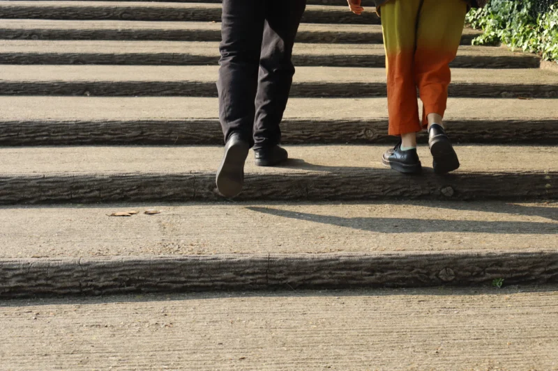 Camilla · 6000 x 4000 Cette photographie met en scène les **pieds de deux personnes** marchant sur un passage piéton. Le **sujet principal** est les chaussures, dont une noire et une verte.La **composition** est simple et épurée, avec un **cadrage serré** centré sur les pieds. Les **lignes droites** du sol et des marches guident le regard. La **lumière** naturelle, probablement en journée, crée des ombres marquées, ajoutant du contraste.Les **couleurs dominantes** sont les tons neutres du béton et les couleurs vives des chaussures. Le style est **minimaliste et documentaire**, évoquant une **ambiance quotidienne et urbaine**.
