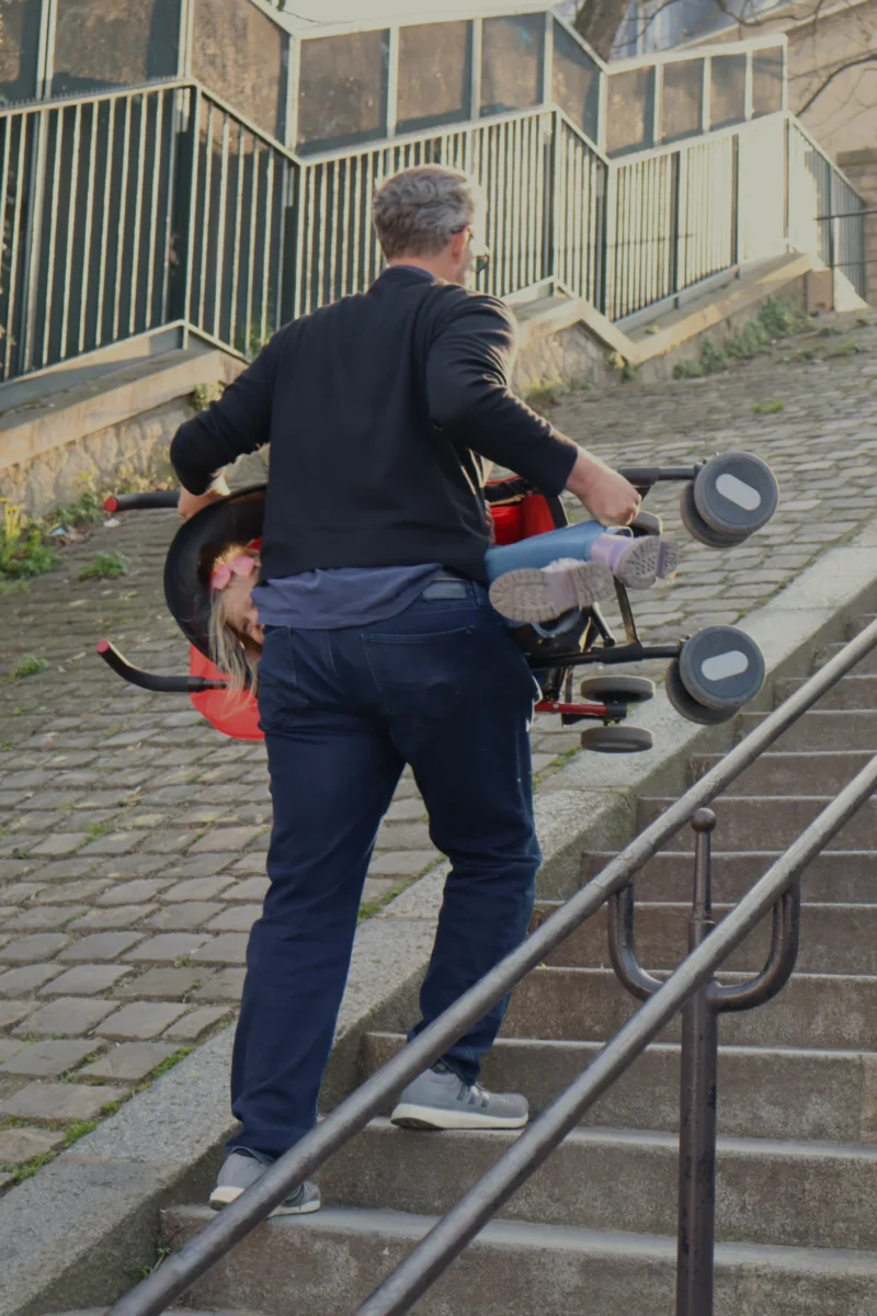 Camilla · 4000 x 6000 Cette photographie montre une personne âgée poussant un enfant dans un landau sur un escalier mécanique extérieur. Le sujet principal est le duo en mouvement.La composition est dynamique, avec un cadrage serré sur les personnages et l’escalier métallique. La lumière semble naturelle, légèrement tamisée, créant des ombres douces. Les couleurs dominantes sont les tons neutres (gris de l’escalier, bleu du vêtement) avec des touches de rouge et noir dans le landau.Le style est documentaire et spontané, capturant un moment du quotidien. L’ambiance est à la fois calme et active, évoquant une scène urbaine paisible.