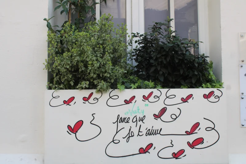 Roberta · 21/03/2026 · 6000 x 4000 Cette photographie montre une fenêtre avec des graffitis en forme de cœurs et d'oiseaux rouges stylisés. Le sujet principal est l'inscription manuscrite en bas, partiellement illisible, avec des motifs décoratifs.La composition est simple et symétrique, centrée sur les motifs. Le cadrage est serré, mettant en valeur les détails des dessins. La lumière semble naturelle, éclairant uniformément la scène. Les couleurs dominantes sont les verts des plantes et les rouges des graffitis, créant un contraste vibrant.Le style est épuré et artistique, évoquant une ambiance à la fois poétique et un peu mélancolique.