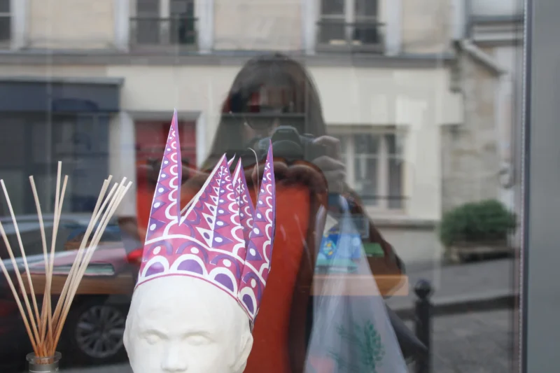Roberta · 21/03/2026 · 6000 x 4000 Cette photographie met en scène une tête sculptée stylisée, probablement en céramique, avec des traits minimalistes et une expression neutre. Le cadrage est serré, centré sur l'objet, avec un flou d'arrière-plan suggérant une rue urbaine.Les couleurs dominantes sont les tons pastel (rose, bleu) des éléments colorés accrochés autour, ainsi que le rouge vif d’un tissu. La lumière semble naturelle, douce, créant des contrastes subtils. L’ambiance est à la fois artistique et mystérieuse, mêlant tradition et modernité dans un cadre urbain. Le style évoque un mélange de street art et d’artisanat.