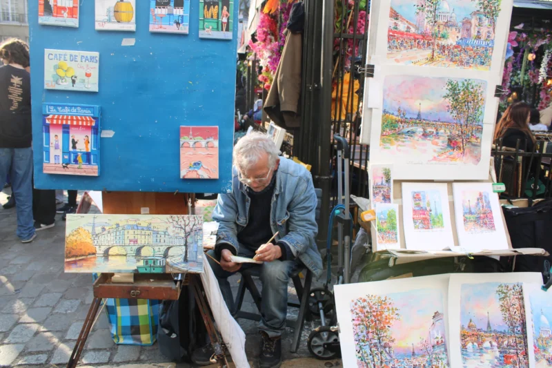 Roberta · 21/03/2026 · 6000 x 4000 Cette photographie montre un vieil homme assis en fauteuil roulant, dessinant ou signant des œuvres. Il est entouré d’une multitude de tableaux colorés, exposés sur des chevalets et des supports.La composition est dynamique, avec un cadrage serré sur le sujet principal, mettant en valeur son activité. La lumière semble naturelle, éclairant les œuvres et créant des contrastes doux. Les couleurs dominantes sont vives et variées, allant des tons chauds aux teintes pastel.L’ambiance est artistique et chaleureuse, évoquant un marché ou une foire aux arts. Le style est réaliste et spontané, reflétant un moment authentique de création.