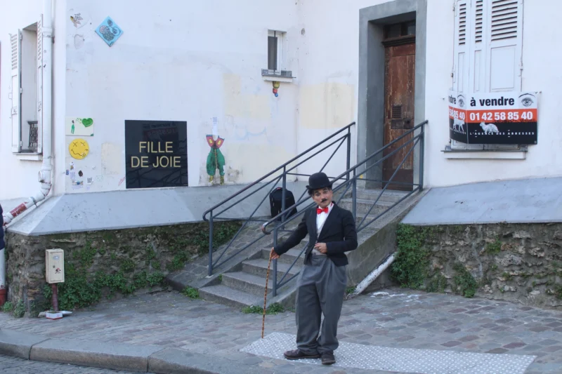 Roberta · 21/03/2026 · 6000 x 4000 Cette photographie montre un homme en costume noir et chapeau, jouant de l'accordéon devant un immeuble. Le sujet principal est cet homme, situé au premier plan.La composition met en valeur les escaliers menant à une porte d’entrée, avec des enseignes ("Fille de joie" et "À vendre"). Le cadrage est serré sur l’artiste, avec un arrière-plan légèrement flou.La lumière est naturelle, douce, et les couleurs dominantes sont les tons neutres (gris, beige) avec des touches de vert et de bleu. L’ambiance est à la fois mélancolique et pittoresque, évoquant un quartier historique et artistique.