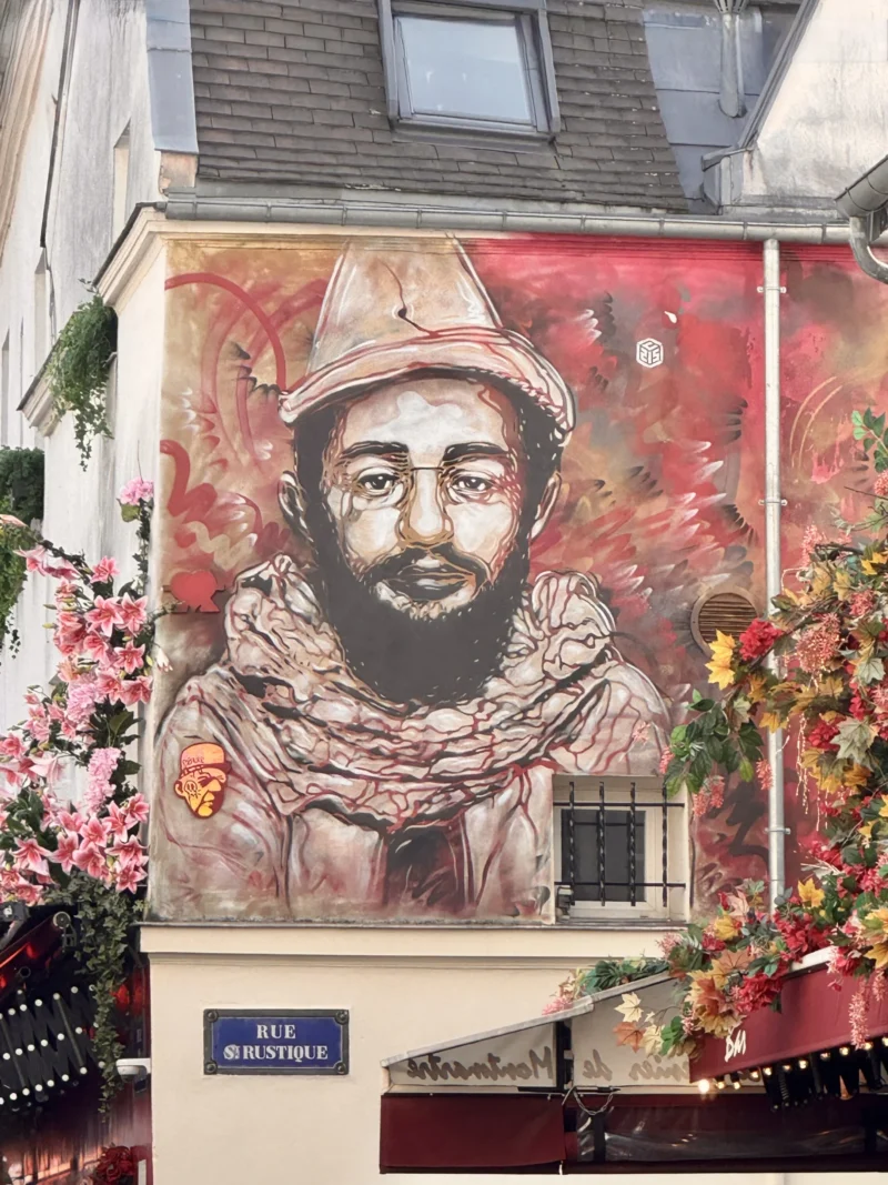 Heike · 21/03/2026 · 3024 x 4032 Cette photographie montre un mural représentant un homme portant un béret et un manteau, avec une barbe et un regard intense. La composition est verticale, centrée sur le visage du personnage. Le cadrage est serré, mettant en valeur le détail du portrait.Les couleurs dominantes sont des tons chauds : rouge, orange et brun, avec des touches de noir et blanc. La lumière semble naturelle, éclairant le mural de manière homogène. Le style est réaliste et graphique, évoquant une ambiance à la fois artistique et historique, renforcée par les fleurs colorées en arrière-plan.