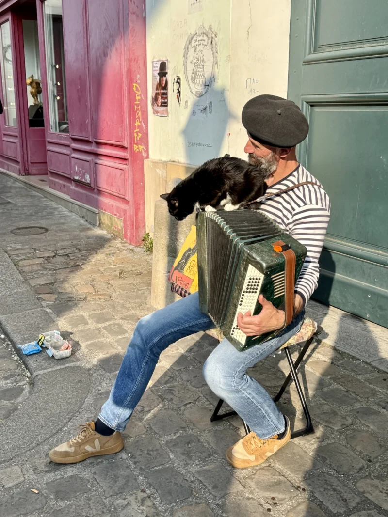 Heike · 21/03/2026 · 4284 x 5712 Cette photographie montre un homme assis sur une marche d’escalier, jouant de l’accordéon. Son chien noir est posé sur l’instrument. La composition est serrée, mettant en valeur les deux sujets principaux.Le cadrage est légèrement bas, créant une ambiance intime. La lumière est naturelle et douce, probablement en journée. Les couleurs dominantes sont vives : les tons pastel des murs (rose, bleu), les couleurs neutres de l’homme (gris, bleu) et les touches chaudes de l’accordéon (bois, métal).Le style est documentaire et spontané, évoquant une scène de rue authentique et chaleureuse.