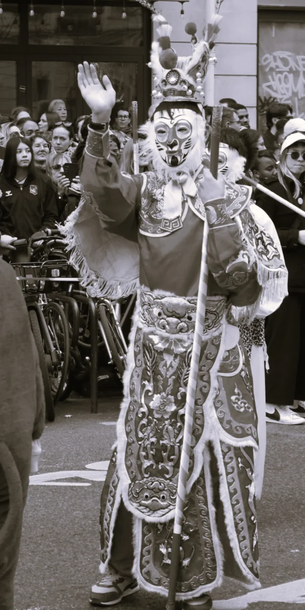 Sophie · 1895 x 3790 Cette photographie en noir et blanc montre un personnage vêtu d’un costume traditionnel richement ornementé, probablement lié à une célébration culturelle ou un défilé. Le sujet principal, au centre, semble être un danseur ou un participant en costume traditionnel asiatique, levant les bras.La composition est dynamique, avec une foule en arrière-plan. Le cadrage est serré sur le personnage, mettant en valeur ses détails vestimentaires. La lumière est naturelle, contrastée, accentuant les contours. L’ambiance est festive et théâtrale, évoquant une tradition vivante.