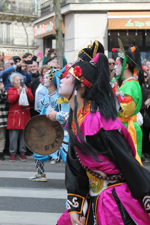 Sophie · 2848 x 4272 Cette photographie capture une scène festive avec pour sujet principal une personne déguisée en masque traditionnel, vêtue de couleurs vives. Elle porte un costume noir et rose, avec des accessoires colorés et un tambourin.La composition montre une rue animée, avec plusieurs participants en arrière-plan. Le cadrage est serré sur le personnage principal, mais inclut aussi des éléments du contexte urbain.Les couleurs dominantes sont vives : rose, noir, vert et jaune. La lumière semble naturelle, probablement en journée. L’ambiance est joyeuse et festive, évoquant une célébration ou un carnaval.