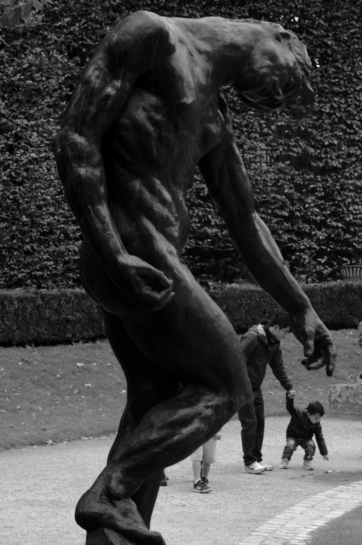 Thomas · 05/10/2025 · 2238 x 3365 Cette photographie en noir et blanc met en scène une statue humaine en mouvement, comme si elle courait ou sautait. Le sujet principal, une silhouette dynamique et stylisée, domine la composition.Le cadrage est serré sur la statue, avec un arrière-plan flou montrant deux personnes et un mur texturé. La lumière semble naturelle, créant des contrastes marqués et des ombres profondes. L'absence de couleurs accentue les jeux de lumière et de texture.Le style est artistique et expressif, évoquant une ambiance à la fois dramatique et poétique, presque cinétique.