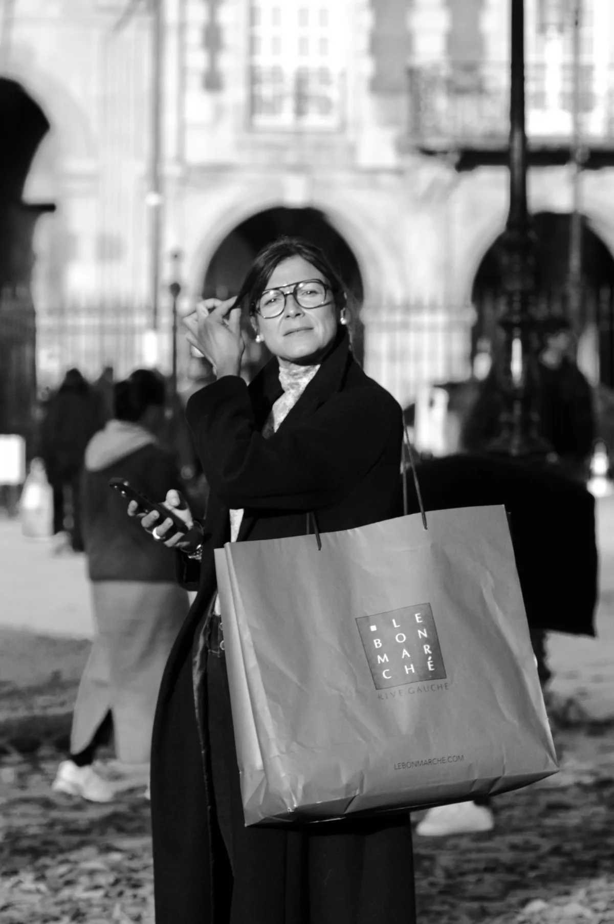 Thomas · 01/11/2025 · 2724 x 4096 Cette photographie en noir et blanc met en scène une femme comme sujet principal, vêtue d’un manteau long et tenant un grand sac à main. Elle semble marcher dans une rue urbaine.La composition est simple et centrée, avec un cadrage serré sur la femme, légèrement décentré. L’arrière-plan montre des bâtiments et des passants floutés, créant une profondeur de champ.La lumière est douce, avec des contrastes modérés, accentuant les textures des vêtements et des détails architecturaux. L’ambiance est sobre et intemporelle, renforcée par le monochrome, évoquant une atmosphère calme et mélancolique.