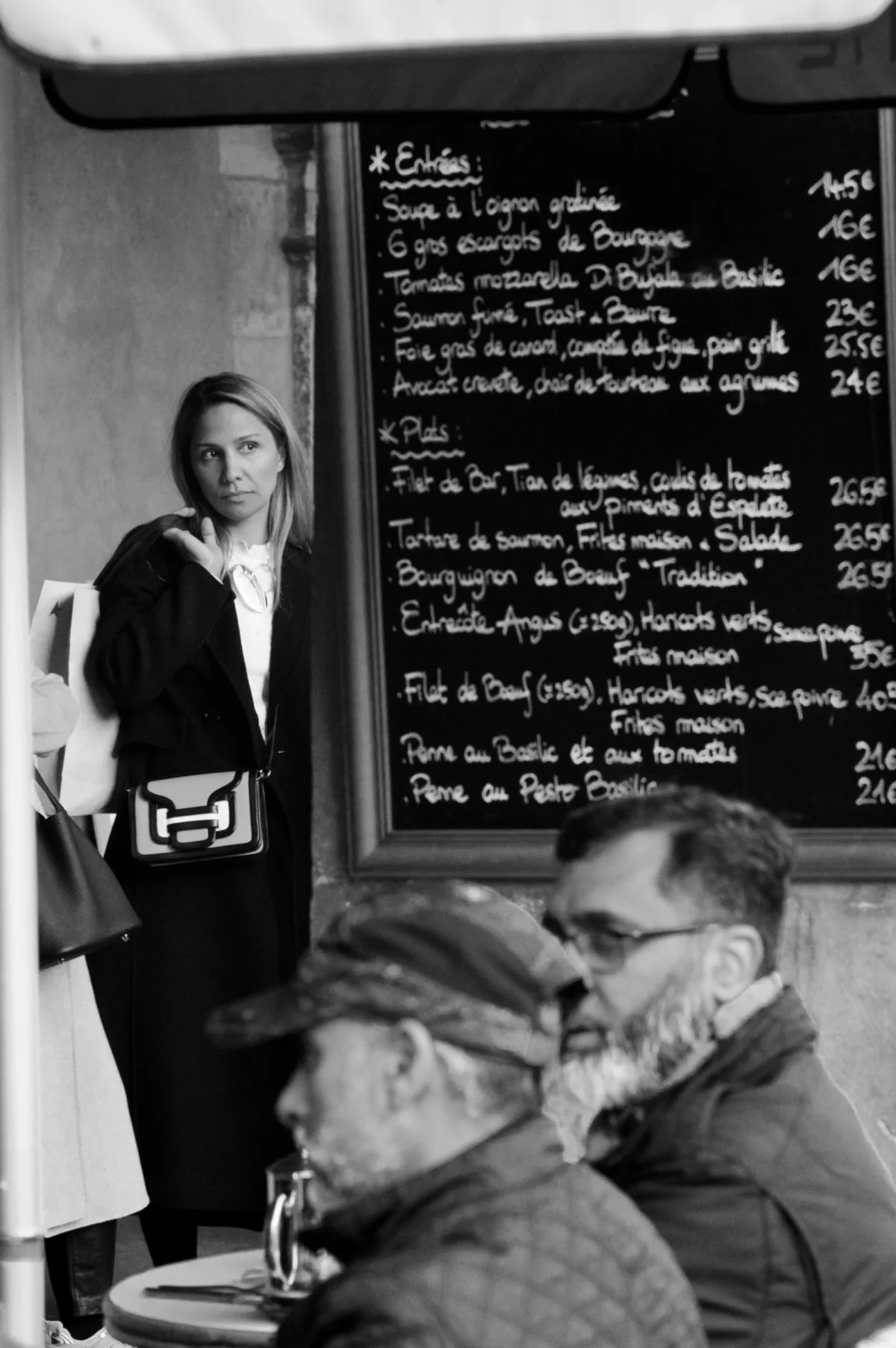 Thomas · 01/11/2025 · 2724 x 4096 Cette photographie en noir et blanc capture une scène intimiste dans un restaurant. Le sujet principal est une femme debout, de dos, près d’un tableau de menu, observant deux hommes assis à une table. La composition joue sur les contrastes entre l’espace intérieur et les regards. Le cadrage serré met en valeur les expressions et les détails des visages. La lumière douce, probablement naturelle, crée des ombres douces et accentue l’ambiance chaleureuse et légèrement mélancolique. Les tons gris dominent, renforçant le côté rétro et intemporel. Le style évoque une esthétique documentaire, typique des clichés des années 1960-1970.