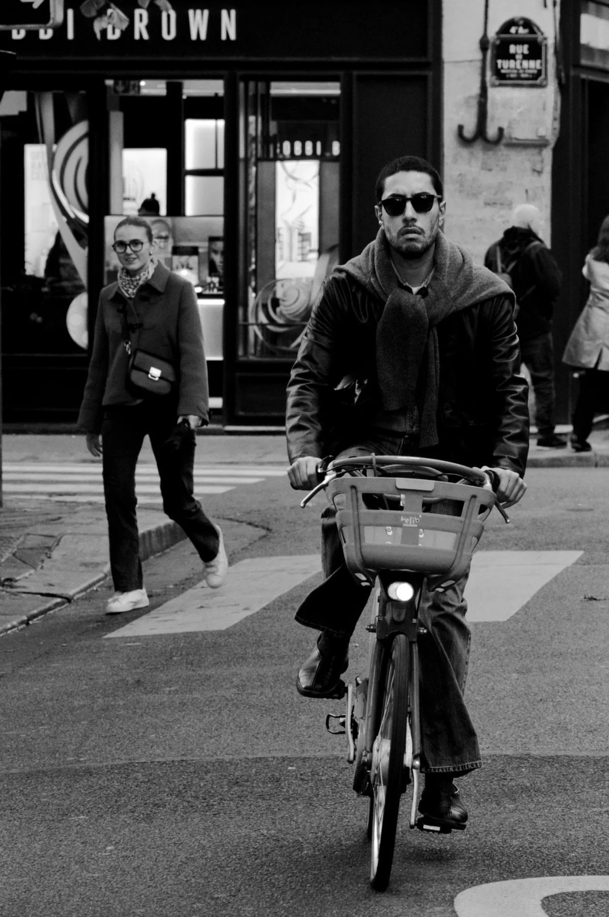 Thomas · 01/11/2025 · 2724 x 4096 Cette photographie en noir et blanc met en scène un homme comme sujet principal, assis sur un vélo avec un panier, vêtu d'une veste longue et de lunettes. La composition est dynamique, avec un cadrage serré sur le cycliste, tandis qu'une femme en arrière-plan ajoute de la profondeur.La lumière est naturelle, contrastée, mettant en valeur les formes et les textures. L'ambiance est urbaine et quotidienne, renforcée par les détails architecturaux et les passants. Le style est documentaire, capturant un instant authentique de la vie citadine. Les tons dominants sont les gris et noirs, typiques du noir et blanc.