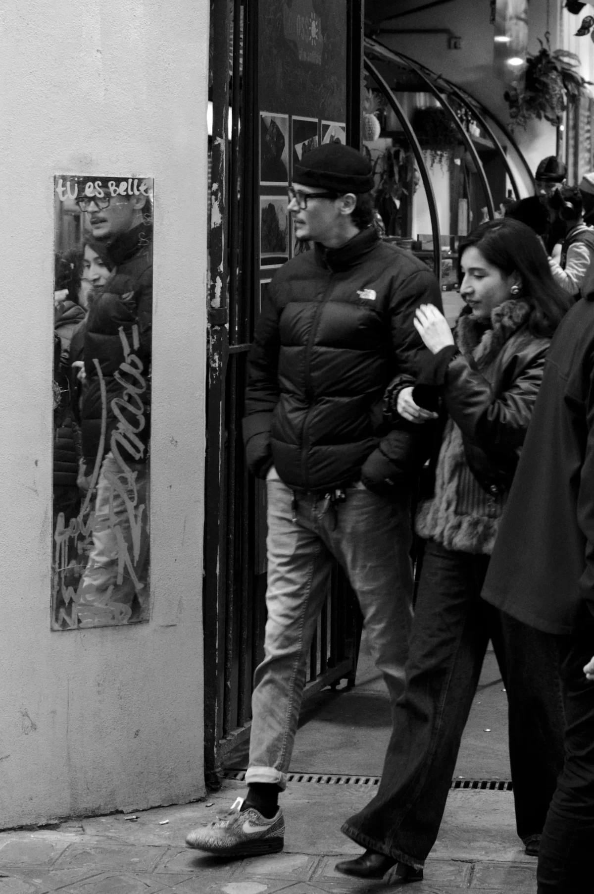 Thomas · 01/11/2025 · 2724 x 4096 Cette photographie en noir et blanc capture deux personnes en extérieur, près d’une entrée. Le sujet principal semble être un homme obèse, vêtu d’un manteau et d’un bonnet, discutant avec une femme en manteau et écharpe. La composition est serrée, centrée sur eux, avec un cadrage en plan moyen.La lumière est douce, probablement artificielle et tamisée, créant des contrastes modérés. L’ambiance est urbaine et nocturne, évoquant une scène intime et discrète. Le style est documentaire, réaliste, mettant l’accent sur les détails humains et l’environnement immédiat.
