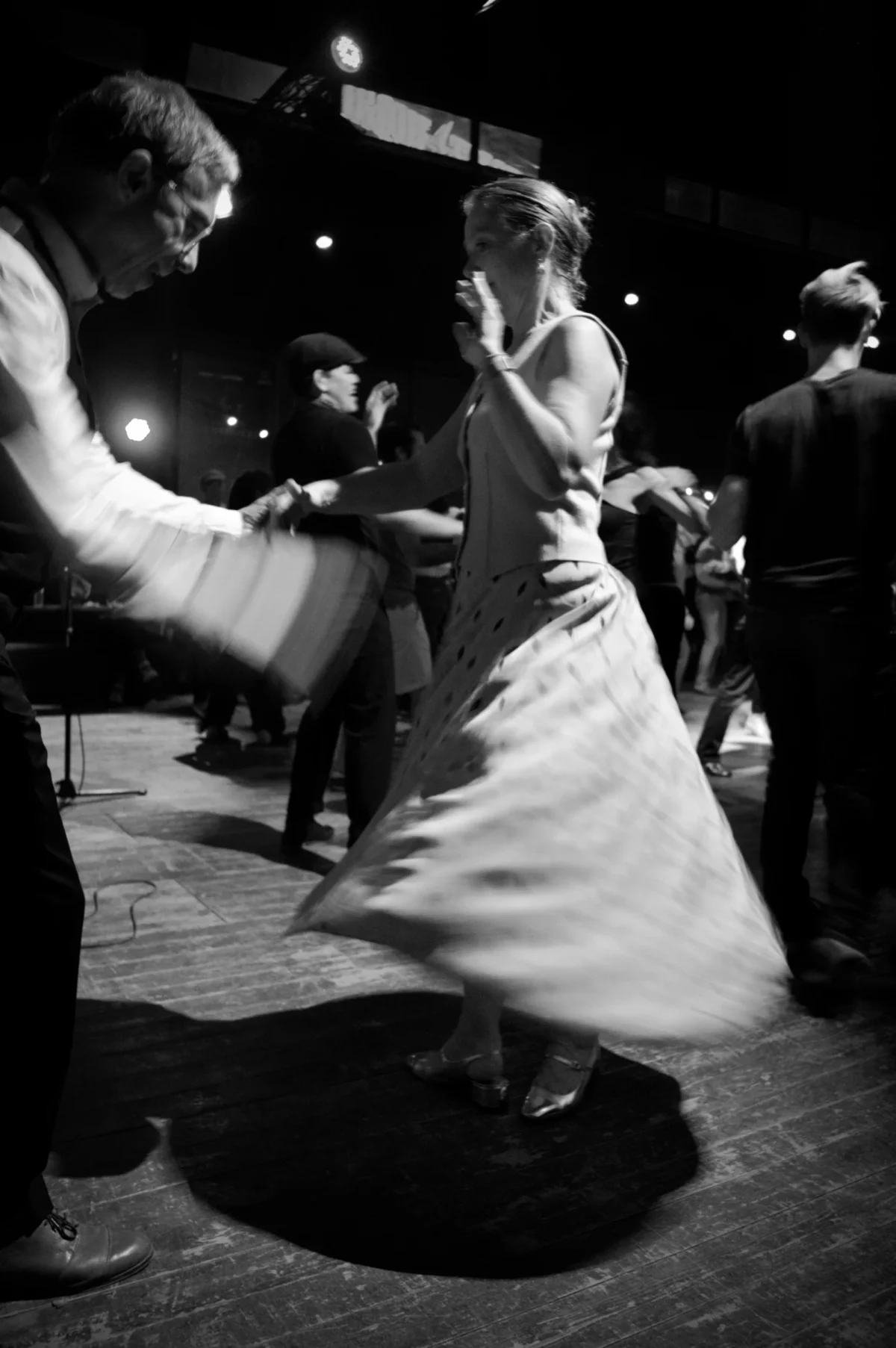 Thomas · 09/11/2025 · 2724 x 4096 Cette photographie en noir et blanc capture un couple en train de danser, probablement un tango. Le sujet principal est la femme en robe longue, en mouvement dynamique, tandis que son partenaire la guide. La composition est serrée, centrée sur eux, avec un cadrage qui isole leur interaction. La lumière est tamisée, créant une ambiance intimiste et dramatique. Les contrastes accentuent les ombres et les mouvements. Le style rappelle le photojournalisme ou documentaire, évoquant une scène authentique et passionnée dans un cadre nocturne et animé.
