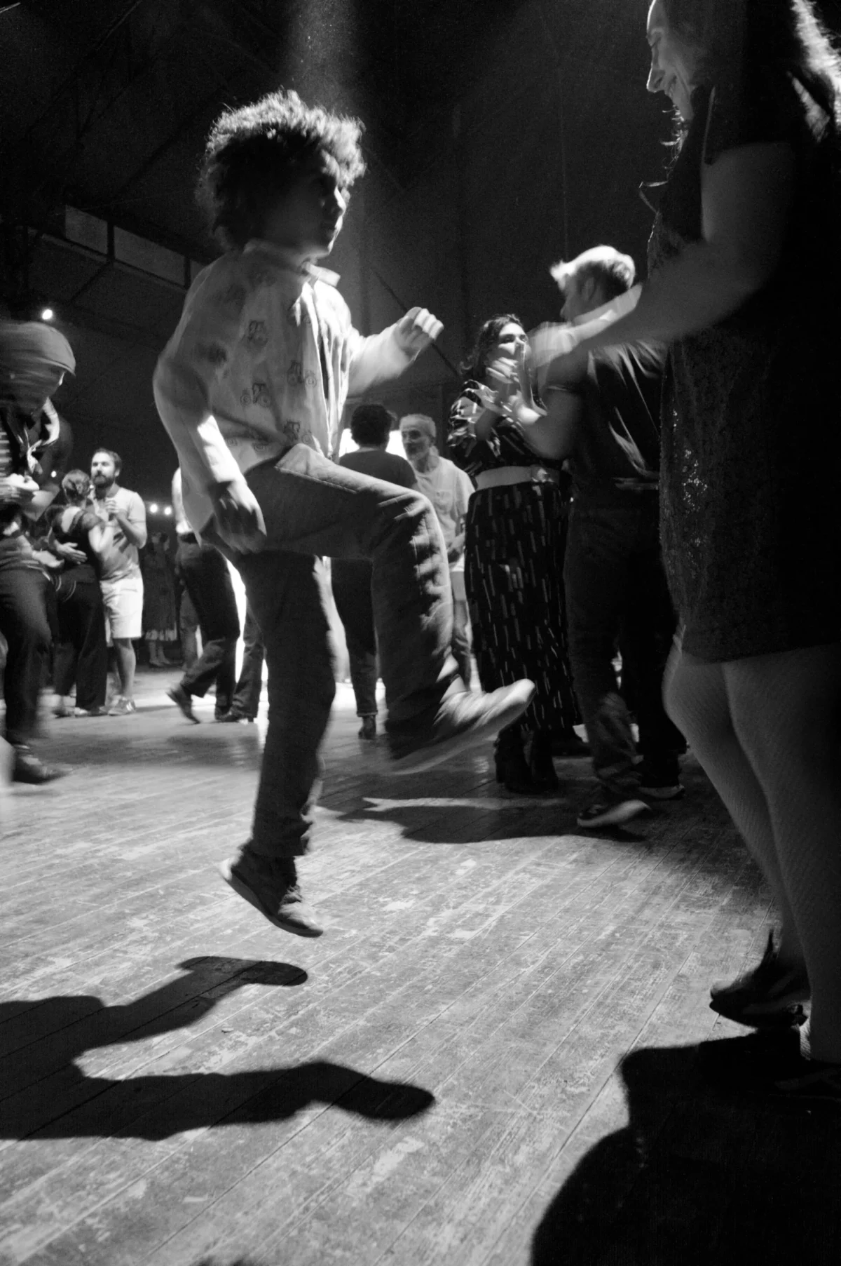 Thomas · 09/11/2025 · 2724 x 4096 Cette photographie en noir et blanc capture une scène de danse dans un lieu bondé. Le sujet principal est un homme en mouvement, au centre, vêtu d’une chemise à carreaux. La composition est dynamique, avec des silhouettes floues en arrière-plan, suggérant une foule en action. Le cadrage serré met l’accent sur le danseur et son énergie.La lumière est tamisée, créant des contrastes marqués et des ombres portées, typiques d’un éclairage scénique. L’ambiance est festive et intense, évoquant une soirée animée, peut-être dans un club ou une salle de concert. Le style est documentaire et spontané, capturant l’instant avec authenticité.