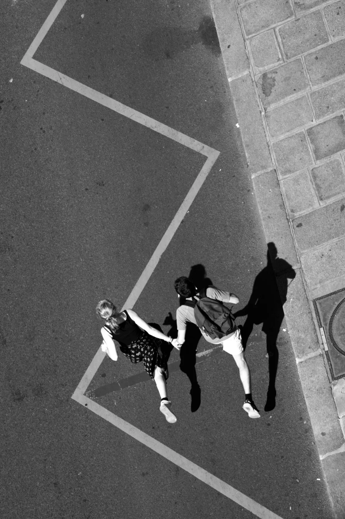 Thomas · 01/05/2025 · 2724 x 4096 Cette photographie en noir et blanc montre deux personnes traversant un passage piéton depuis une vue aérienne. Le sujet principal est une femme tenant un chien en laisse, suivie d’un homme.La composition est épurée, avec une ligne droite du trottoir et un cadrage serré. La lumière semble naturelle, sans ombres marquées, créant un contraste doux.Les tons dominants sont les gris et noirs, renforçant une ambiance sobre et intemporelle. Le style est documentaire et minimaliste, évoquant une scène du quotidien, sobre et dépouillée.