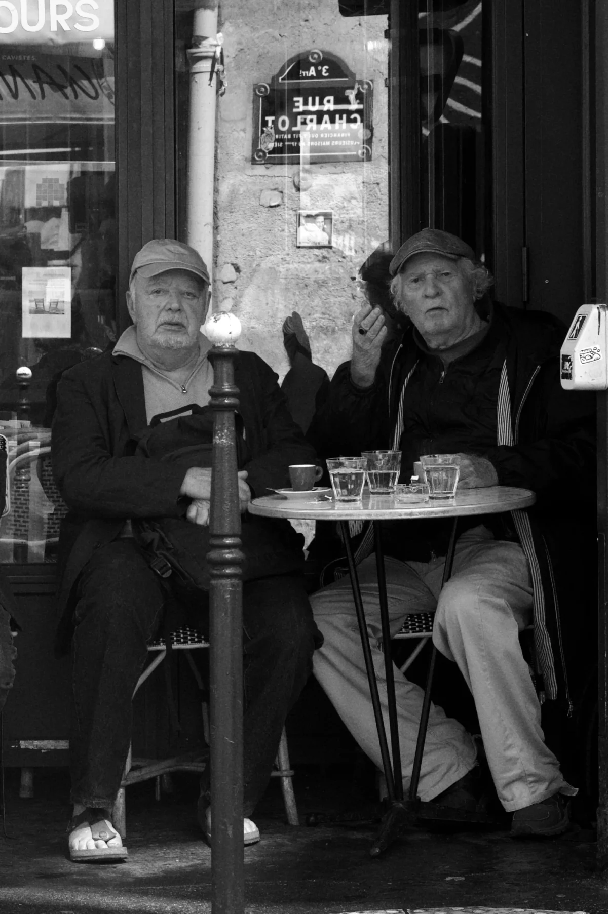 Thomas · 20/07/2025 · 1851 x 2783 Cette photographie en noir et blanc montre deux personnes âgées assises à une table de café en extérieur. Le sujet principal est leur interaction, l’une gesticulant comme pour parler, l’autre écoutant.La composition est serrée, centrée sur eux, avec un cadrage en plan moyen. La lumière est douce, probablement naturelle, créant des contrastes modérés. L’ambiance est intimiste et chaleureuse, évoquant une scène de vie quotidienne et détendue. Le style est documentaire, capturant un moment authentique et spontané. Les tons gris dominent, renforçant l’aspect rétro et intemporel de l’image.