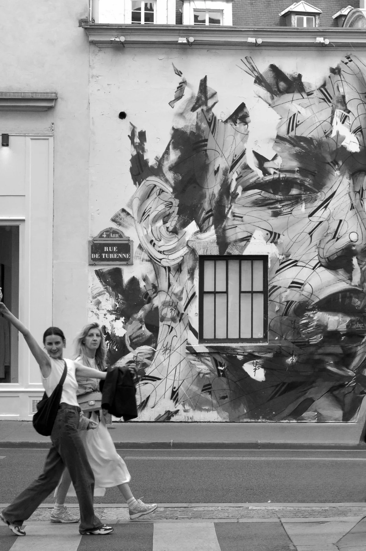 Thomas · 02/08/2025 · 2725 x 4096 Cette photographie en noir et blanc met en scène une femme marchant devant un mur mural. Le sujet principal est cette femme, vêtue d’une robe légère, tenant un objet à la main.La composition joue sur le contraste entre le mouvement de la femme et le dynamisme du mural abstrait derrière elle, représentant des formes tourbillonnantes. Le cadrage est serré, centré sur la femme et le mur.La lumière est naturelle, douce, sans ombres marquées. Les tons dominants sont les gris et noirs, accentuant les contrastes.Le style est documentaire et artistique, avec une ambiance à la fois urbaine et poétique.