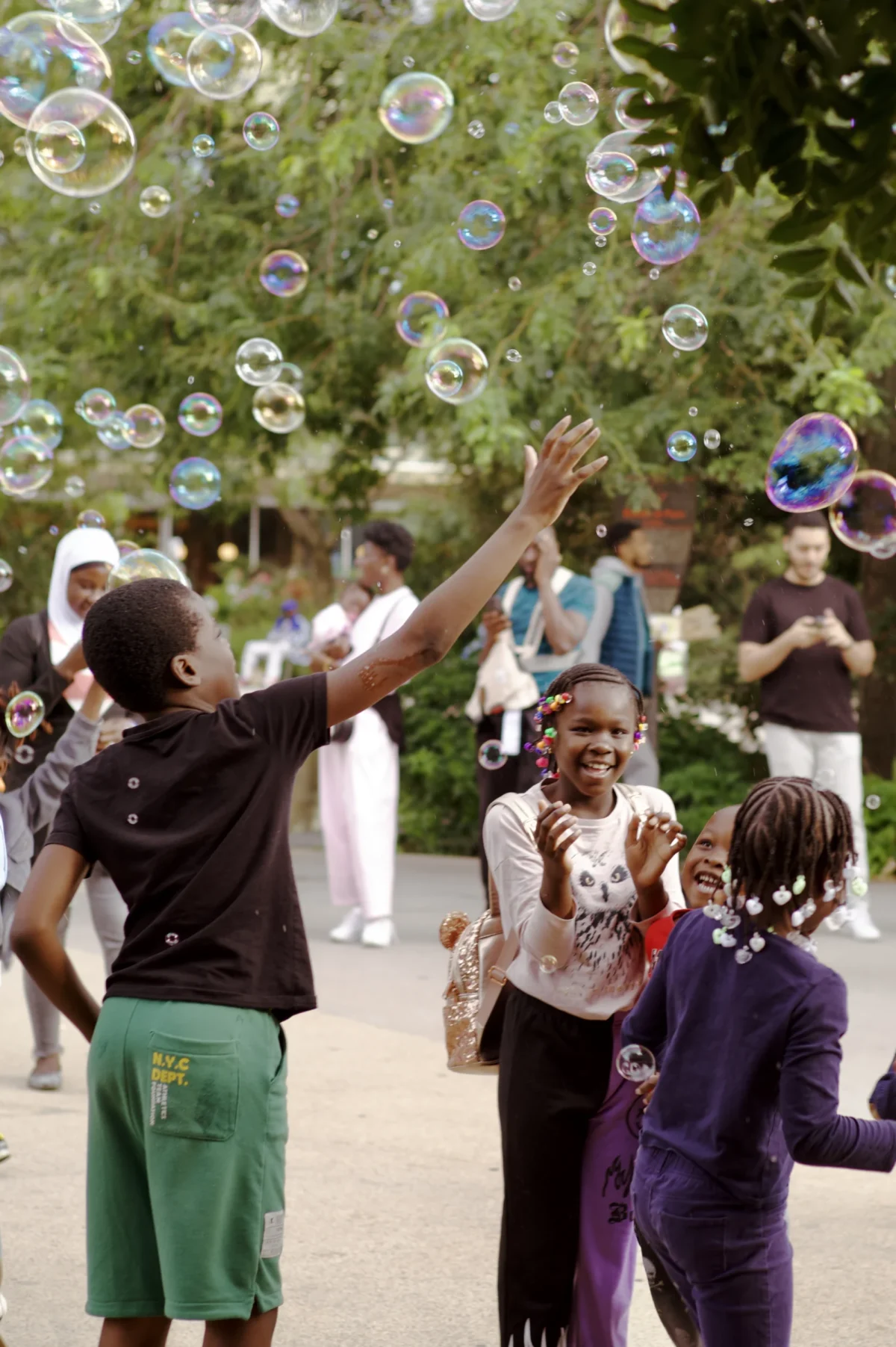 Thomas · 02/08/2025 · 2725 x 4096 Cette photographie capture un groupe d’enfants jouant avec des bulles de savon. Le sujet principal est une fillette levant les bras pour attraper une bulle, entourée d’autres enfants en mouvement.La composition est dynamique, avec un cadrage serré sur les enfants au premier plan et un arrière-plan légèrement flou montrant des adultes et des arbres. La lumière est naturelle et douce, créant une ambiance joyeuse et ensoleillée. Les couleurs dominantes sont vives : verts des arbres, tons pastel des vêtements des enfants, et transparences bleutées des bulles. Le style est spontané et naturel, évoquant une scène de détente et de bonheur.